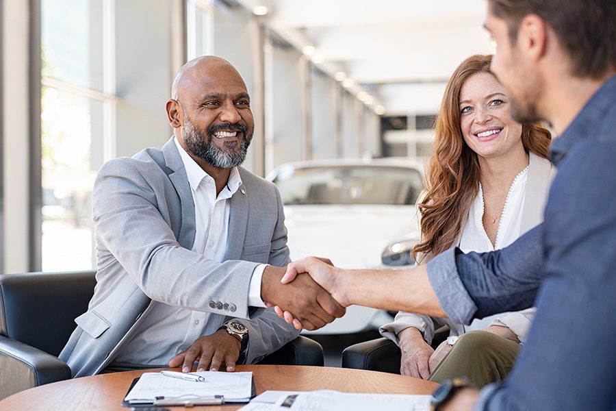 Business Insurance - A Car Salesman is Shaking Hands and Smiling With a Couple While Signing Paperwork at a Dealership