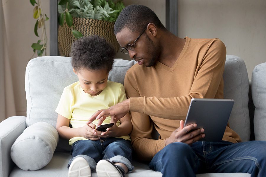 Client Center - A Father and Son are Sitting Together on a Sofa While Holding a Tablet and Smart Phone at Home